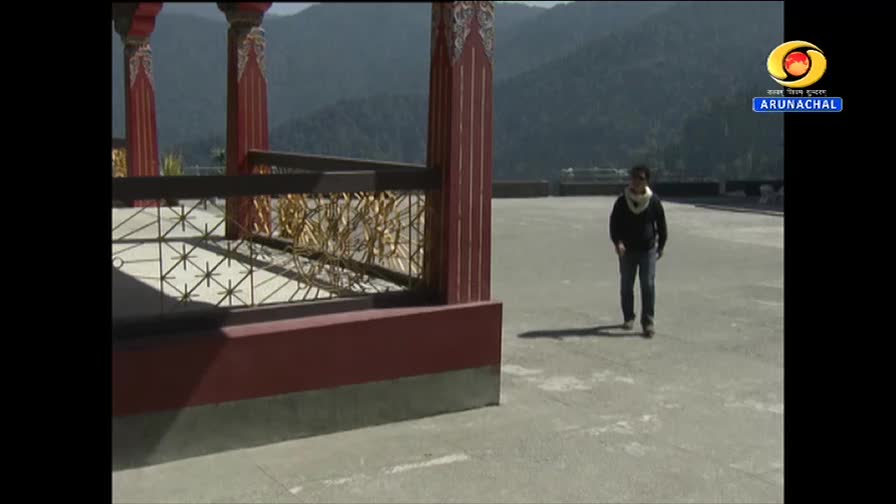 A lone figure walks across an open plaza, the DD Arun Prabha logo visible in the corner. Mountains rise in the background, their slopes covered in trees.