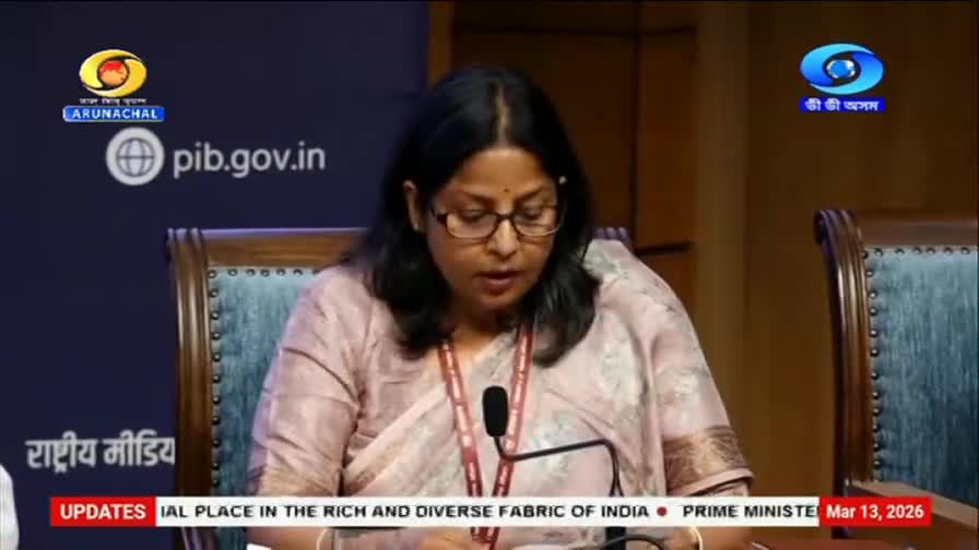 A woman in a light pink sari speaks into a microphone, her glasses perched on her nose. The DD Arun Prabha logo appears on a screen behind her.