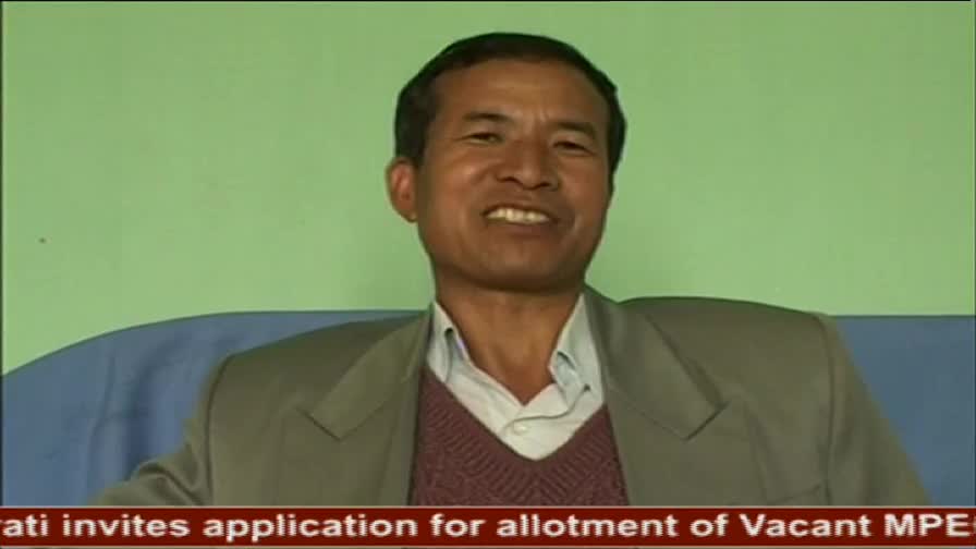 A man with a warm smile sits on a blue couch, his face framed by a light green wall. He wears a grey blazer over a maroon v-neck sweater and a light blue collared shirt.