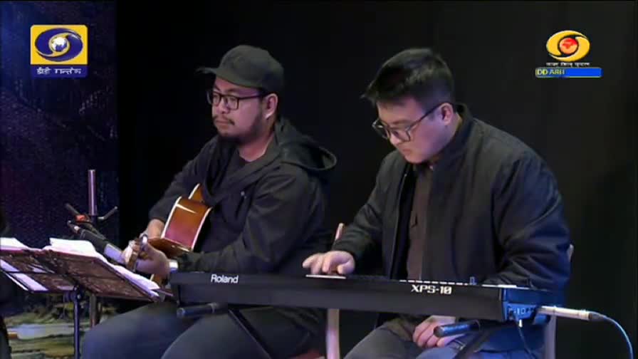 A guitarist, wearing a black cap and glasses, strums his instrument while a keyboardist plays a Roland XPS-18. Both are seated, focused on their music, with the DD Arun Prabha logo visible in the background.