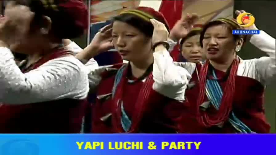 Women in traditional attire adjust headbands as they perform. Their beaded necklaces catch the light, and the DD Arun Prabha logo is visible. Women in traditional attire adjust headbands as they perform. Their beaded necklaces catch the light, and the DD Arun Prabha logo is visible.