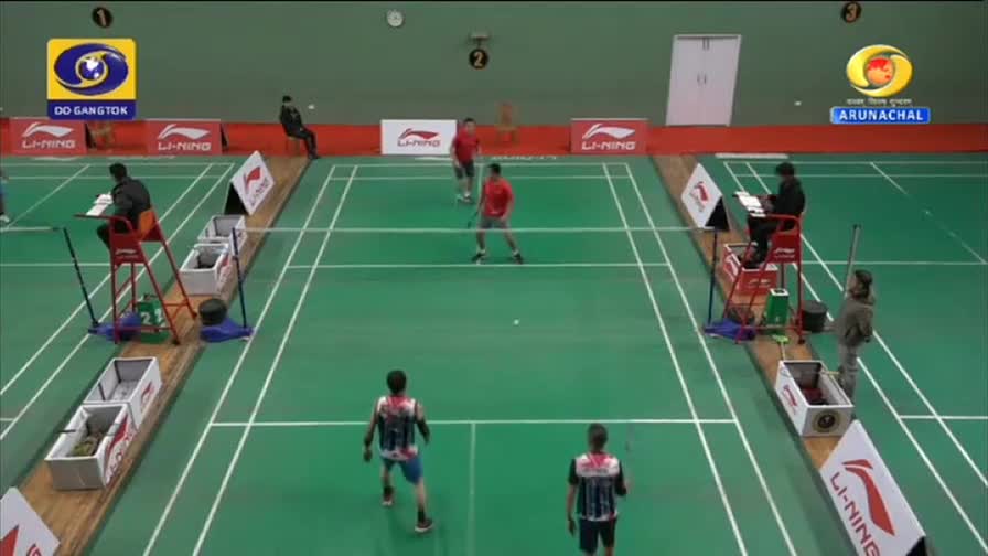 A badminton match is underway, with players in red and patterned shirts moving across the green court. The DD Arun Prabha broadcast shows officials seated high above the court on both sides. A badminton match is underway, with players in red and patterned shirts moving across the green court. The DD Arun Prabha broadcast shows officials seated high above the court on both sides.