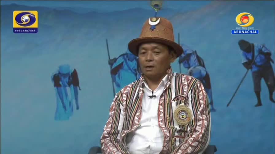 A man in traditional attire speaks, his patterned jacket and distinctive hat catching the light. Behind him, figures in blue, reminiscent of a DD Arun Prabha broadcast, are depicted in a stylized, mountainous landscape. A man in traditional attire speaks, his patterned jacket and distinctive hat catching the light. Behind him, figures in blue, reminiscent of a DD Arun Prabha broadcast, are depicted in a stylized, mountainous landscape.