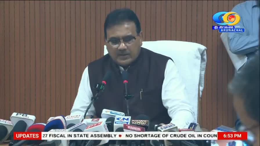 A man in a dark vest over a white shirt speaks into a microphone, surrounded by a cluster of news microphones. The DD Arun Prabha logo appears in the upper right corner. A man in a dark vest over a white shirt speaks into a microphone, surrounded by a cluster of news microphones. The DD Arun Prabha logo appears in the upper right corner.