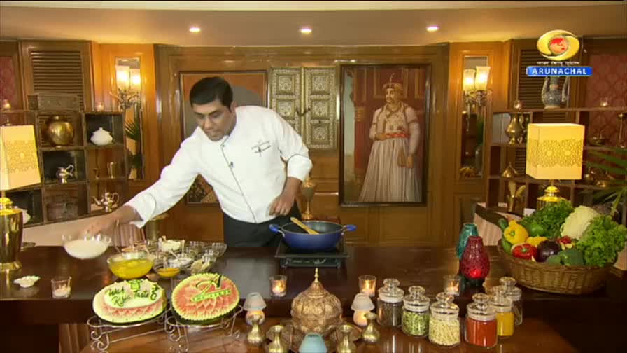 A chef in a white uniform reaches for a bowl of white ingredients. Behind him, a portrait of a regal figure hangs on the wall, and the DD Arun Prabha logo is visible. A chef in a white uniform reaches for a bowl of white ingredients. Behind him, a portrait of a regal figure hangs on the wall, and the DD Arun Prabha logo is visible.