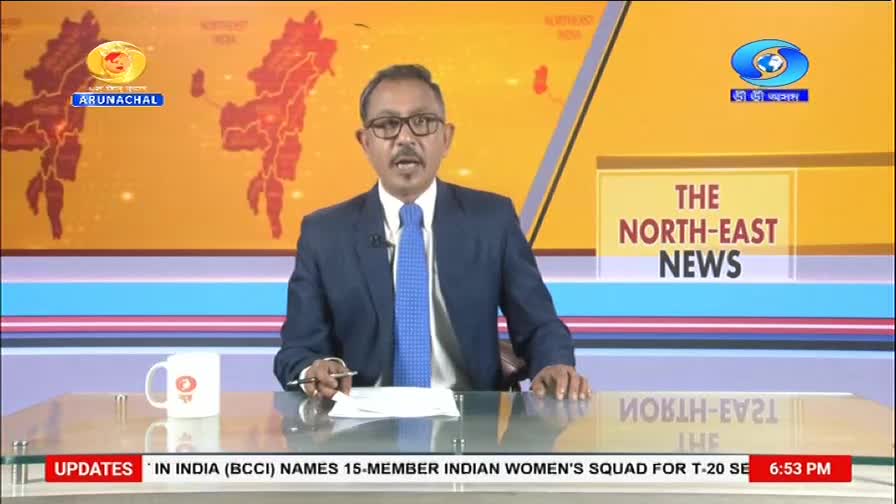 A man in a suit and tie speaks into a microphone, seated at a desk. Behind him, a screen displays maps of India and the text "THE NORTH-EAST NEWS."