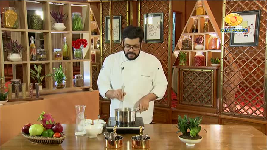 A chef in a white coat stirs a pot on a stovetop, his focus sharp. Behind him, shelves are stocked with jars of spices and decorative items, a scene fitting for a cooking segment on DD Arun Prabha.