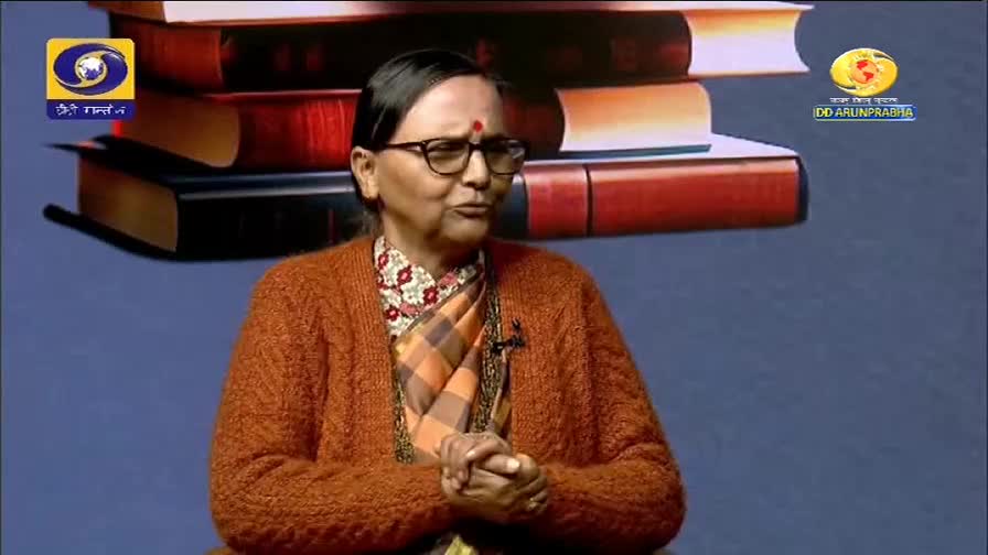 A woman with glasses and a bright orange cardigan speaks, her hands clasped in her lap. Behind her, a stack of books forms a backdrop for the DD Arun Prabha broadcast.