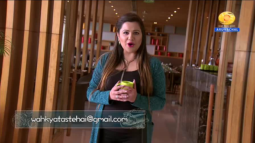 A woman stands holding a bright green mug, her expression animated. Behind her, the DD Arun Prabha logo is visible against a backdrop of warm wood paneling.