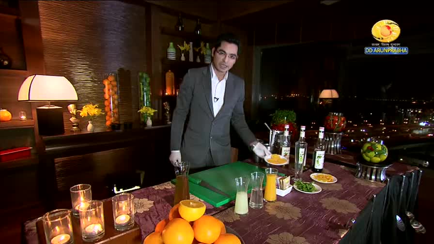 A man in a suit, likely from DD Arun Prabha, presents a plate of sliced oranges. The table before him is laden with fresh fruit, juices, and bottles, set against a backdrop of a city at night.