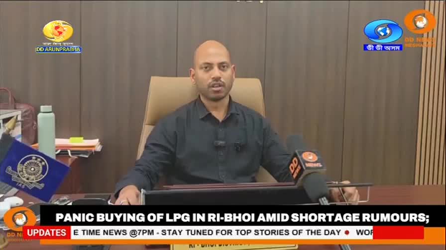 A man in a dark shirt speaks into a microphone at a desk. A banner below him announces "PANIC BUYING OF LPG IN RI-BHOI AMID SHORTAGE RUMOURS."