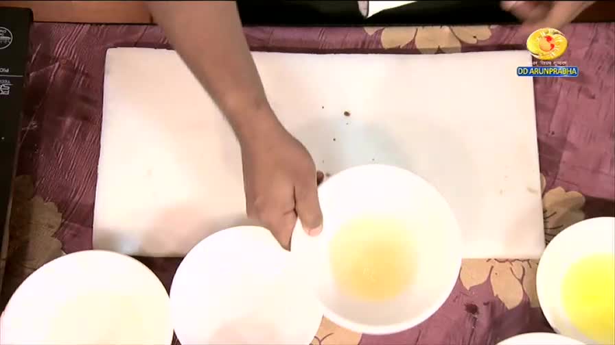 A hand holds a white bowl containing a pale yellow liquid over a large, flat white surface. Another white bowl sits to the left, and a third to the right, both also holding yellow liquid, as seen on DD Arun Prabha.