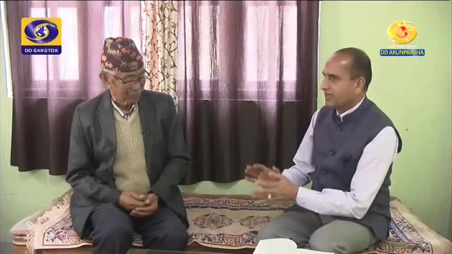 An older gentleman in a traditional cap and a younger man in a vest are seated, engaged in conversation. The DD Arun Prabha logo is visible, indicating a broadcast from India.