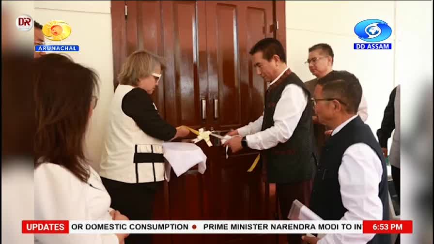 A woman and a man in traditional vests are cutting a yellow ribbon. They stand before a polished wooden door, surrounded by onlookers.