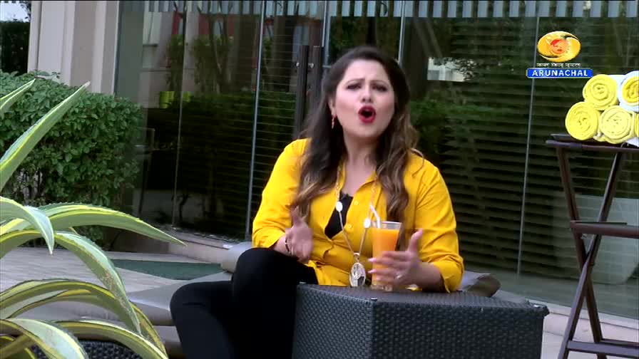 A woman in a bright yellow shirt speaks animatedly, holding a glass of orange juice. In the background, the DD Arun Prabha logo is visible, along with a stack of neatly rolled yellow towels.