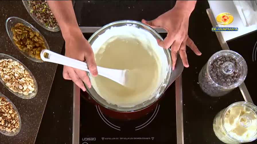 A white spatula stirs a thick, creamy mixture in a glass bowl. Nearby, bowls of nuts and jars of chocolate chips wait their turn.
