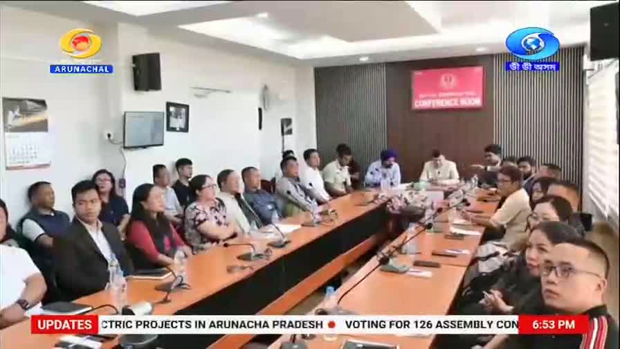 People are gathered around a long conference table, some listening intently, others looking towards the front. A screen above them displays "CONFERENCE ROOM" in red lettering, with the DD Arun Prabha logo visible in the corner.