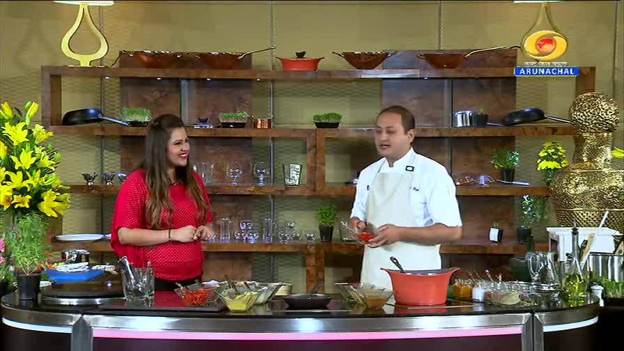 A chef in a white apron holds up a small red ingredient, speaking to a woman in a bright red top. They stand in a studio kitchen, with shelves of cookware and decorative items behind them, likely for a DD Arun Prabha broadcast.
