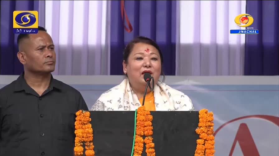 A woman speaks into a microphone, her face illuminated by stage lights, while a stern-faced man in a black shirt stands guard beside her. The backdrop features the DD Arun Prabha logo, suggesting a broadcast from Arunachal Pradesh.