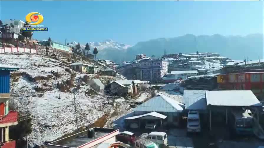 A light dusting of snow covers the hillside village, with several vehicles parked in front of buildings. The DD Arun Prabha logo is visible in the upper left corner.