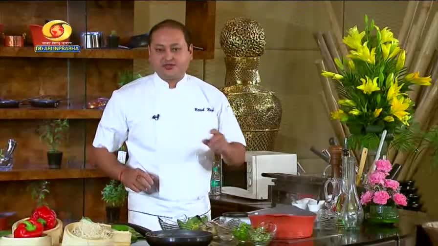 A chef in a white coat gestures as he speaks, with a bright bouquet of yellow lilies behind him. On the counter, red bell peppers sit in a bamboo steamer next to a bowl of noodles, all part of a DD Arun Prabha cooking segment.