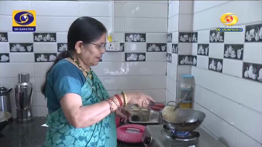 A woman in a teal sari is frying something in a kadai on a gas stove. The DD Arun Prabha logo is visible in the background.