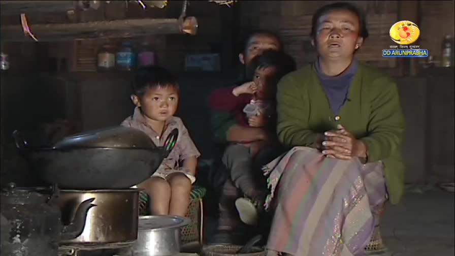 A woman in a green cardigan speaks, her hands clasped, while a young boy watches intently beside a large cooking pot. Behind them, another woman holds a child as they sit in what appears to be a home in India, with the DD Arun Prabha logo visible.