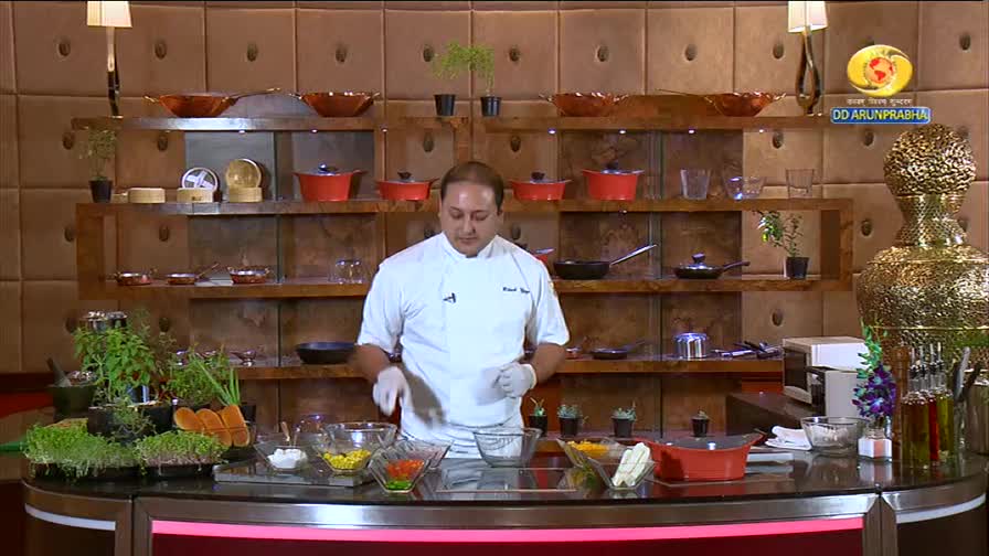 A chef in a white coat, on DD Arun Prabha, stands before a counter filled with fresh ingredients. He's preparing a dish, his gloved hands poised over bowls of colorful vegetables.