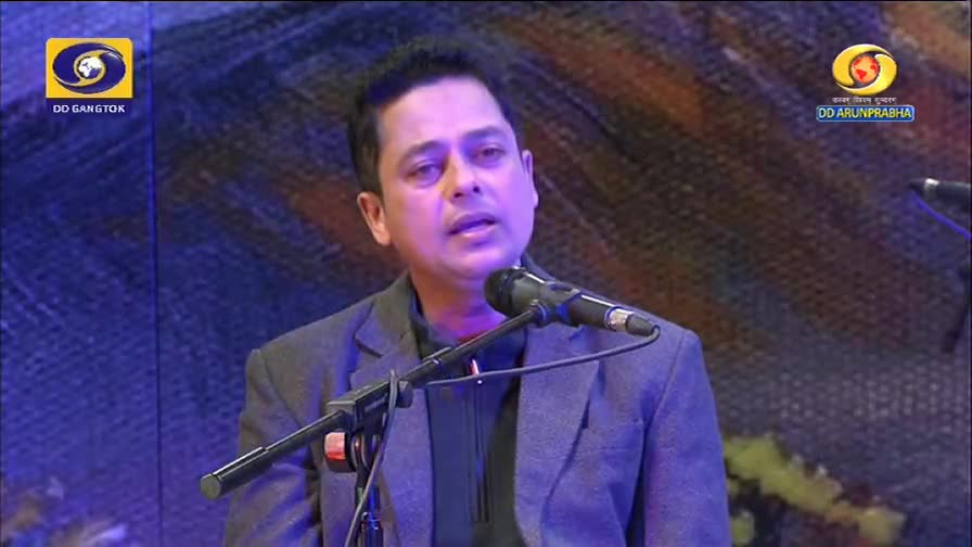 A man in a grey suit speaks into a microphone on a stage. The DD Arun Prabha logo is visible in the background.