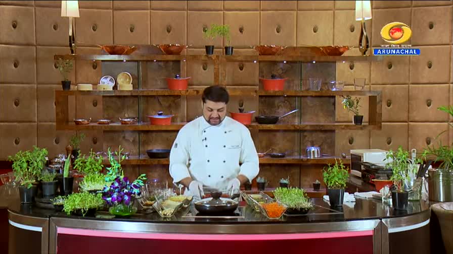 A chef in a white coat stands behind a counter, his hands hovering over a black pot. Fresh green sprouts and vibrant purple orchids are arranged on the counter, with shelves of cookware and decorative items behind him. The DD Arun Prabha logo is visible in the upper right corner.