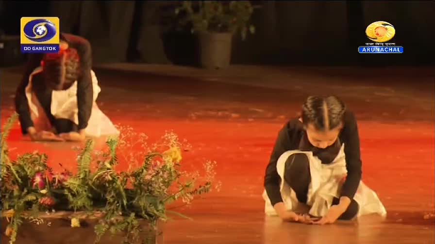 Two young dancers, dressed in black tops and white skirts, are kneeling on the stage. They are performing a synchronized routine, their heads bowed low. The stage lights cast a warm, orange glow across the polished floor.
