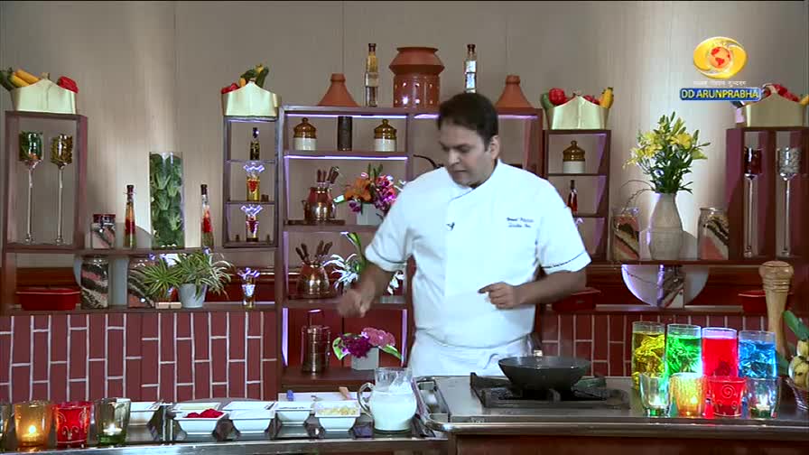 A chef in a white uniform gestures as he stands in front of a stove on the DD Arun Prabha set. Colorful drinks line the counter next to him, reflecting the studio lights.