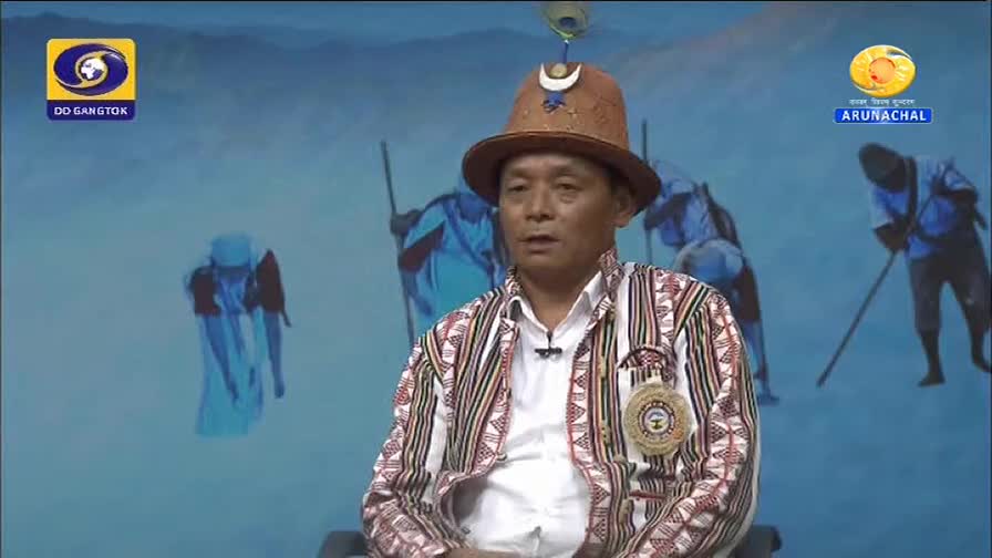 A man in a traditional hat and patterned jacket sits before a backdrop depicting figures in blue, some holding long poles. The DD Arunachal logo is visible on the right.
