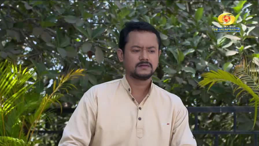 A man in a light beige kurta stands outdoors, his expression thoughtful. Lush green foliage surrounds him, with a glimpse of the DD Arun Prabha logo in the upper right corner.