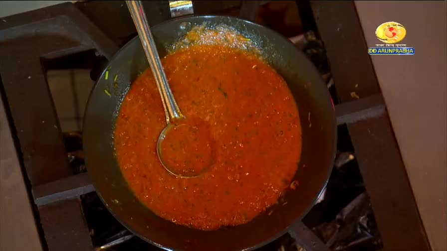 A rich, red curry simmers in a dark pan, steam rising gently. A ladle rests submerged, ready to serve this dish from DD Arun Prabha.