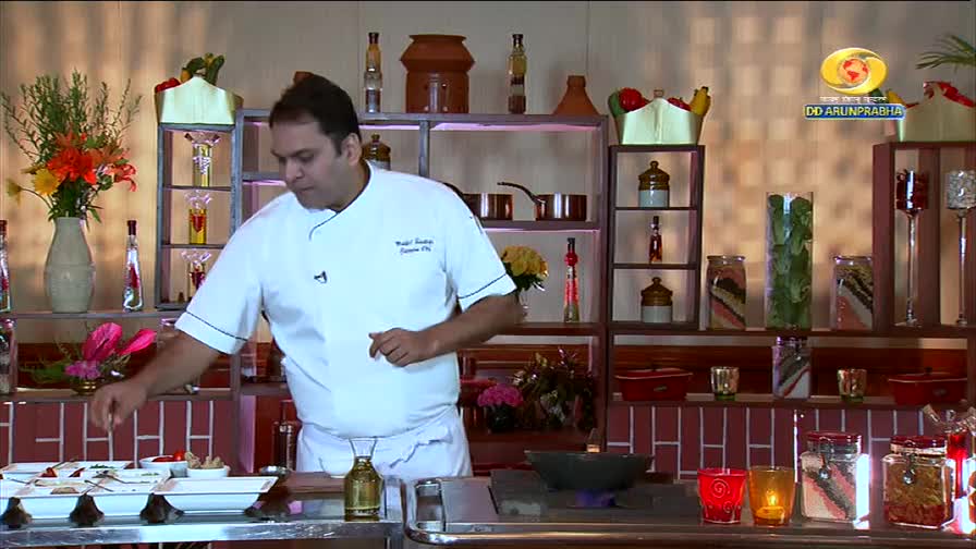 A chef in a white uniform carefully arranges small dishes on a counter. Behind him, shelves display decorative jars and cookware, with the DD Arun Prabha logo visible.