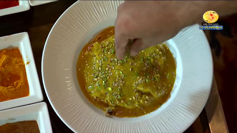 Fingers sprinkle chopped nuts over a bright yellow curry. Nearby, small dishes hold vibrant orange and brown spices, ready for the cooking show on DD Arun Prabha.
