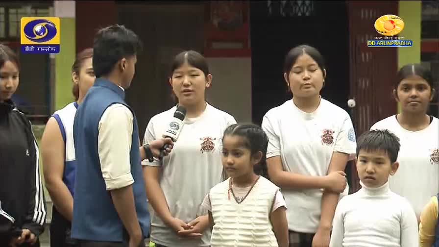 A young man in a blue vest interviews a group of children and young women, their faces turned towards him. The DD Arun Prabha logo is visible in the background, indicating a broadcast from India.