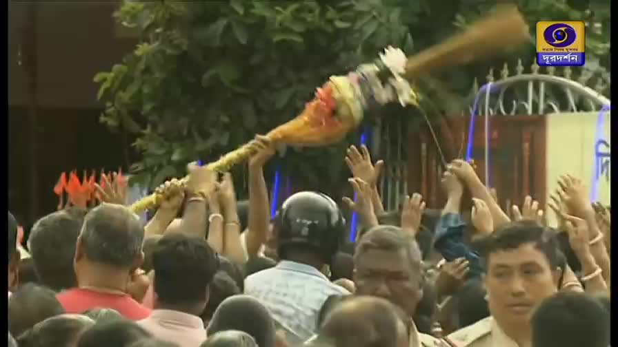 A large, decorated object is held aloft by many hands, its end adorned with flowers and a brush-like element. People reach up, their faces turned toward the object, as the camera angle suggests this is being broadcast on DD Bangla.
