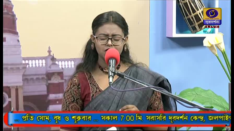 A woman with glasses sings into a red microphone, her eyes closed. Behind her, the DD Bangla logo and a backdrop with a building are visible.
