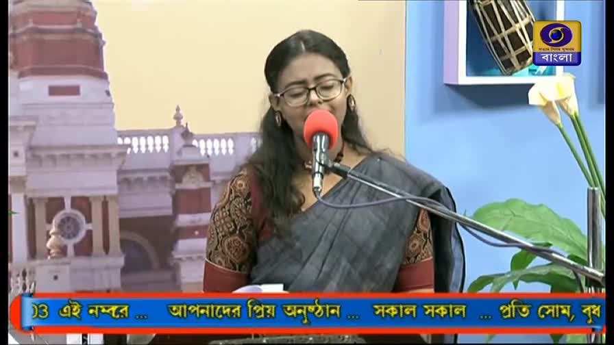 A woman in a gray sari sings into a red microphone, her eyes closed. Behind her, the DD Bangla logo and a backdrop of a building are visible.
