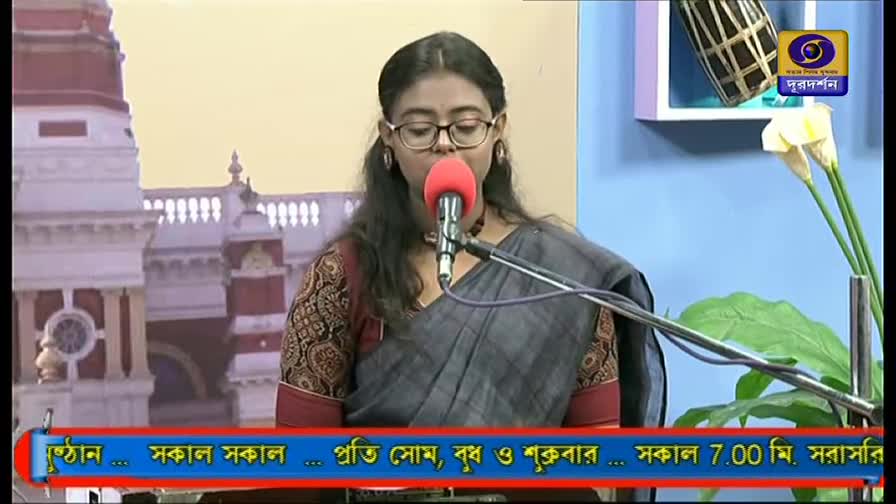 A woman in glasses, wearing a saree, sings into a red microphone. The DD Bangla logo sits in the corner, and the lower third banner announces the show's schedule.
