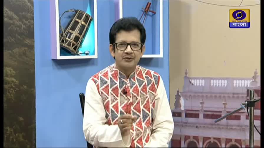 The man in glasses gestures with his right hand while speaking, his patterned vest a splash of color against the blue backdrop. The DD Bangla logo is visible in the corner, and behind him, a shelf displays a drum and a stringed instrument.
