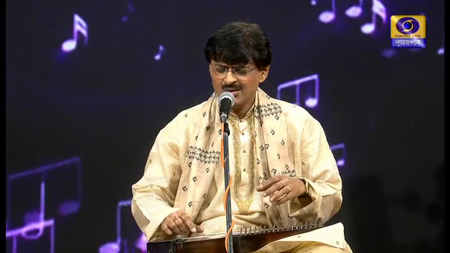 A man in glasses, wearing a cream-colored kurta, sings into a microphone while seated. His fingers pluck at the strings of a small instrument resting on his lap, as the DD Bangla logo subtly glows in the background.
