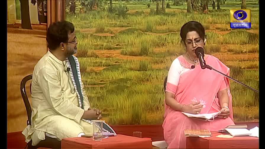 A woman in a pink sari sits at a table, reading from a book, while a man in a kurta listens intently. The DD Bangla logo is visible in the corner, and the backdrop depicts a rural scene.
