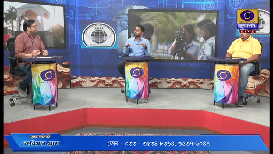 Three men sit behind individual desks, each with a DD Bangla logo, in a brightly lit studio. One man gestures with his hands while the others listen, and a screen behind them shows a scene of people working with cameras.
