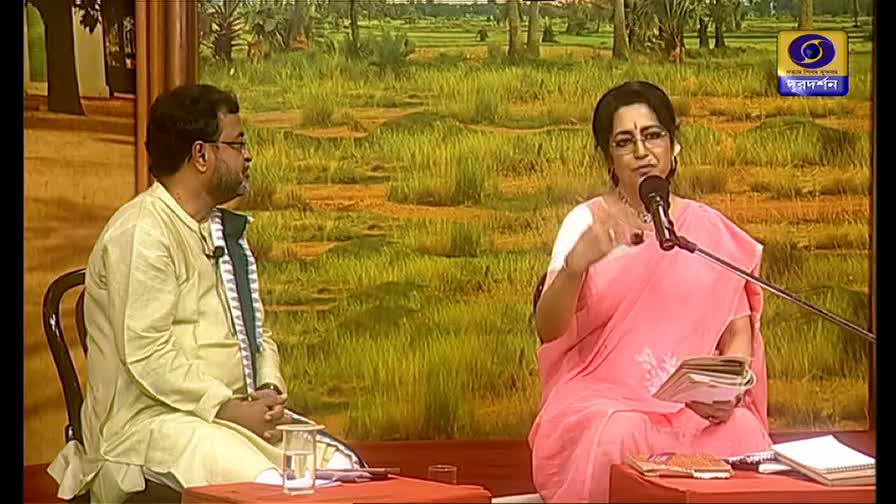A woman in a pink sari gestures emphatically, speaking into a microphone. Beside her, a man in a light yellow kurta listens intently.
