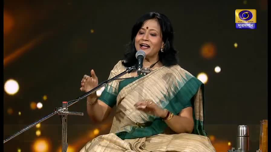 A woman in a sari sings into a microphone, her hands gesturing gracefully. The DD Bangla logo is visible, and the stage is lit with soft, golden light.
