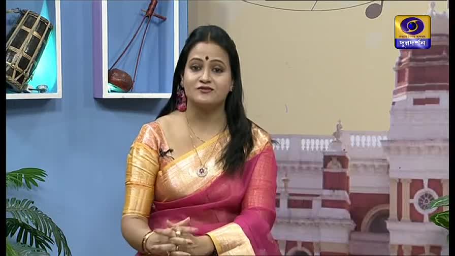 The woman in the pink sari is speaking, her hands clasped in front of her. Behind her, the DD Bangla logo and a backdrop of a building are visible.
