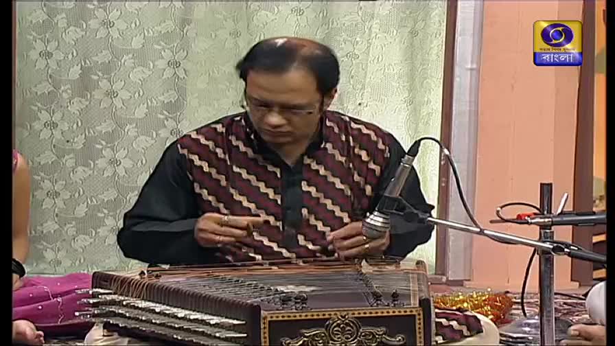 A man in a patterned shirt is playing a stringed instrument, his fingers moving delicately over the strings. The DD Bangla logo is visible in the corner, suggesting a live performance.
